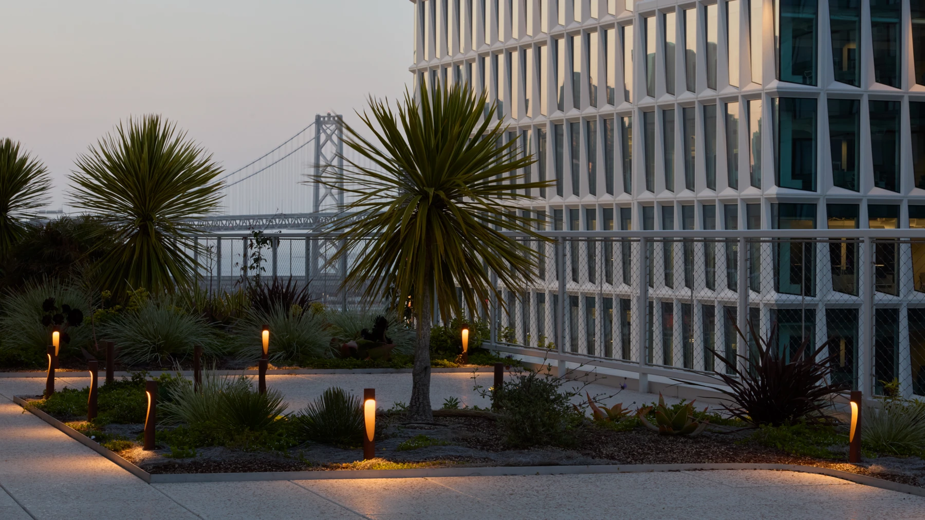 Flindt Bollard lights line a path through a planted urban garden beside a modern glass building. © 2026 Louis Poulsen.