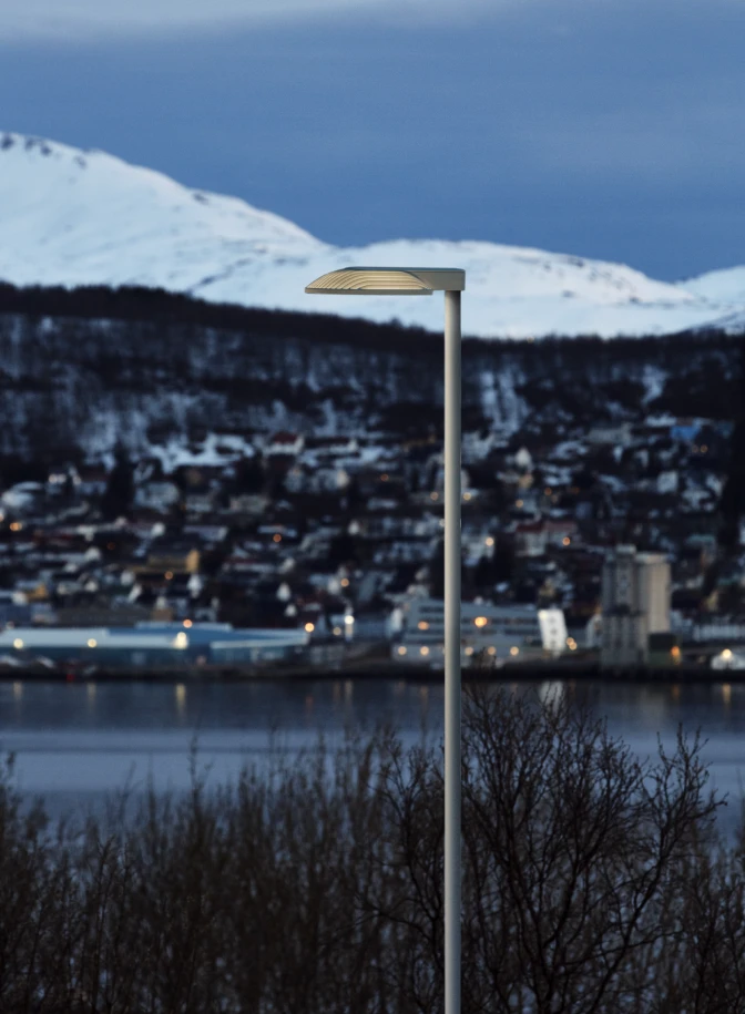 Una luminaria urbana Homann Park ilumina un pueblo nevado junto al agua, con montañas al fondo. © 2026 Louis Poulsen.