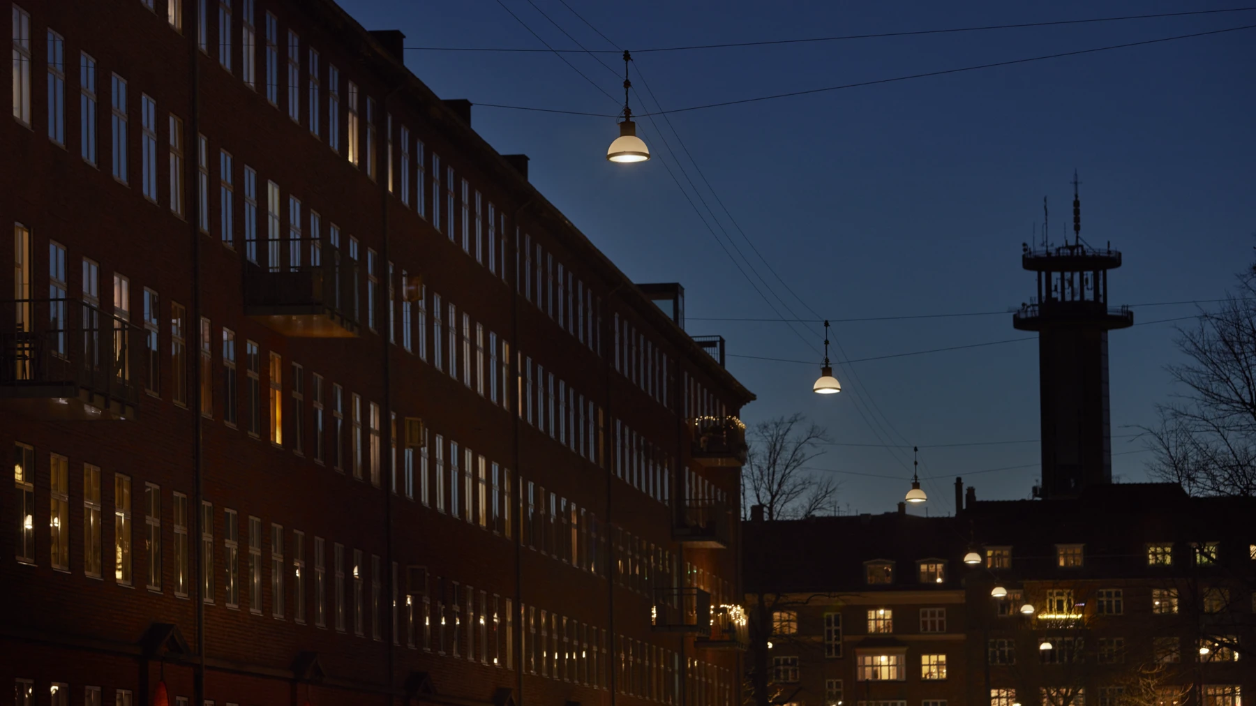 Las luminarias suspendidas Ballerup iluminan una calle residencial entre edificios de ladrillo al anochecer. © 2026 Louis Poulsen.