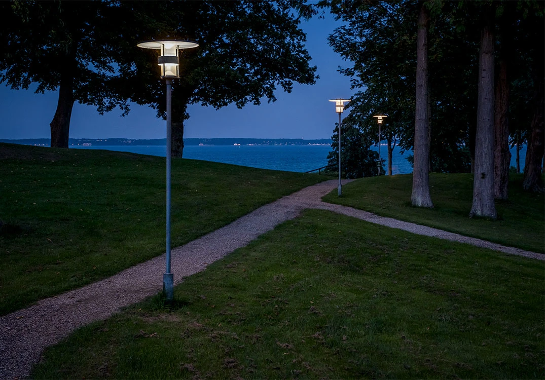 Des Albertslund Maxi Post éclairent un chemin de gravier bordé d’arbres au bord de l’eau, au crépuscule. © 2026 Louis Poulsen.