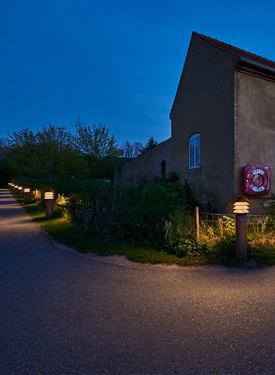 Des bornes Bysted éclairent un chemin bordé de végétation le long d’un bâtiment au crépuscule. © 2026 Louis Poulsen.