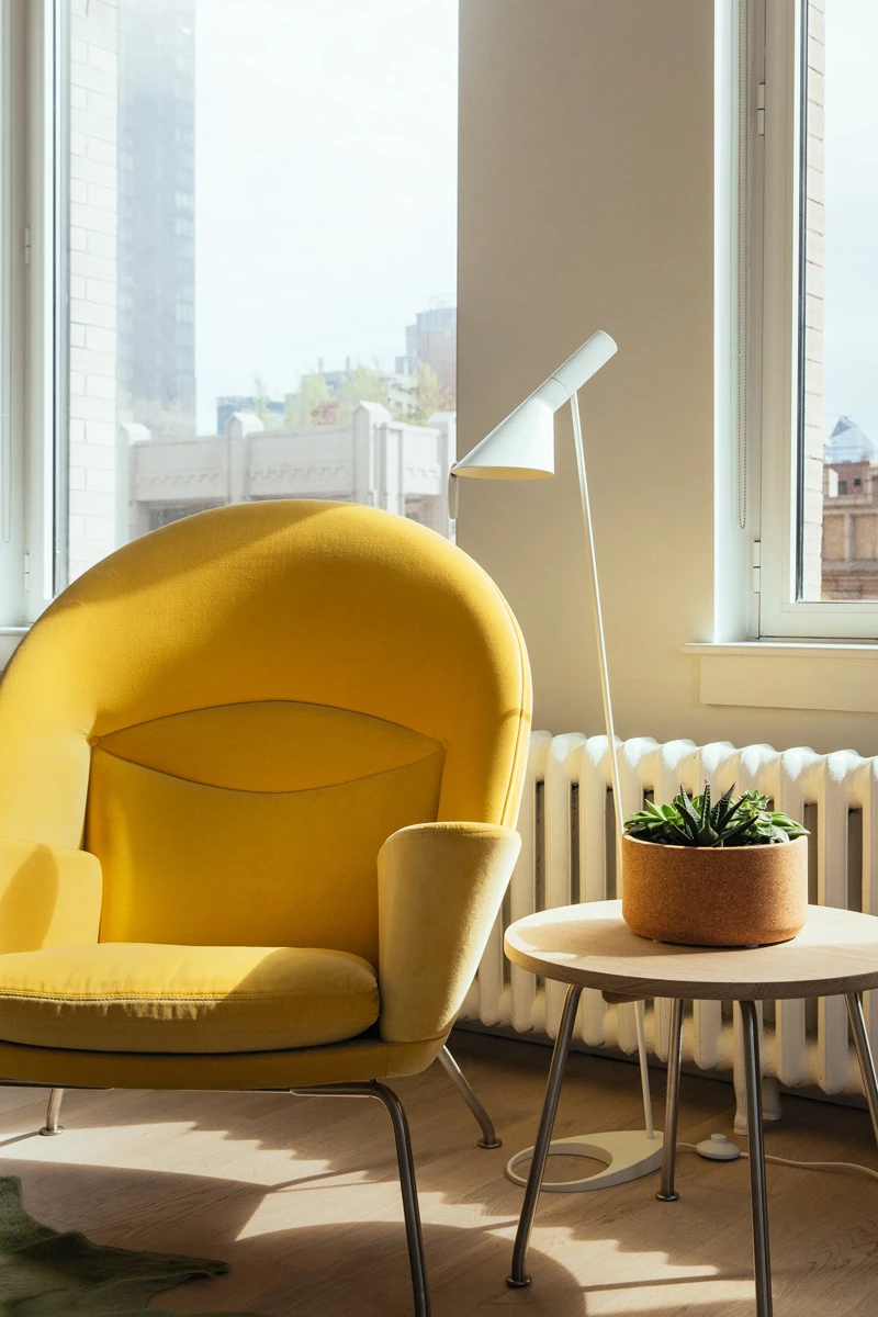 Una AJ Floor Lamp bianca è accanto a una poltrona gialla e a un tavolino vicino a grandi finestre. © 2026 Louis Poulsen.