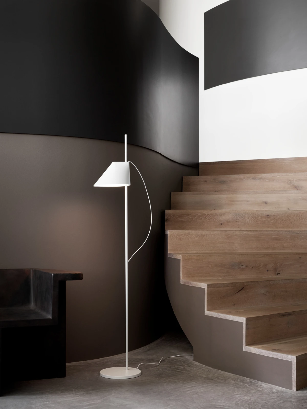 A white Yuh Floor Lamp stands beside a sculptural wooden staircase against dark walls. © 2026 Louis Poulsen.