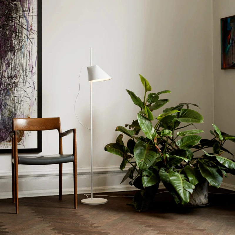 Un lampadaire Yuh blanc se tient près d’une chaise en bois et d’une grande plante verte dans un salon. © 2026 Louis Poulsen.