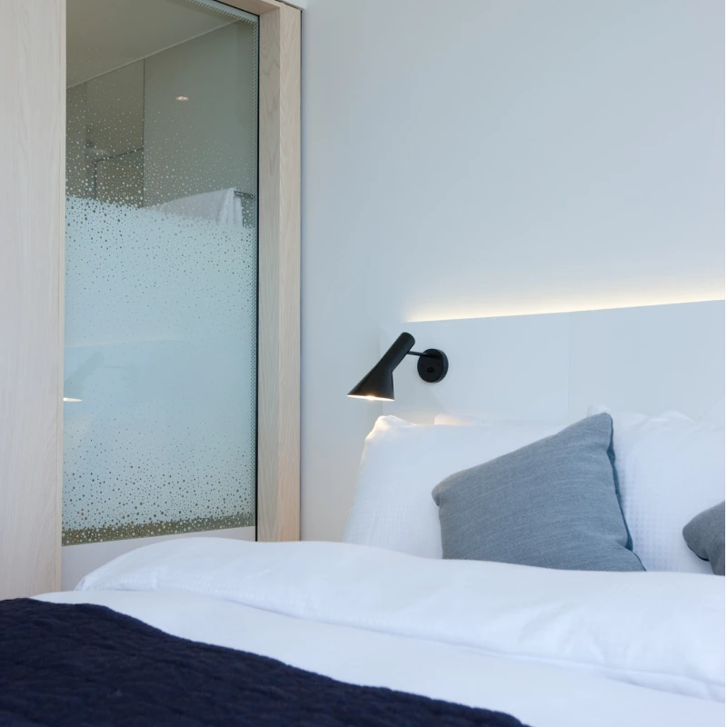 明亮卧室床边的黑色AJ Wall Lamp,搭配白色床品和灰色靠枕。 © 2026 Louis Poulsen.