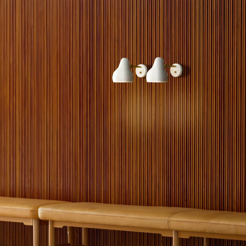 Two white VL 38 Wall Lamps with brass details on a vertical wood-slat wall above a leather bench. © 2026 Louis Poulsen.