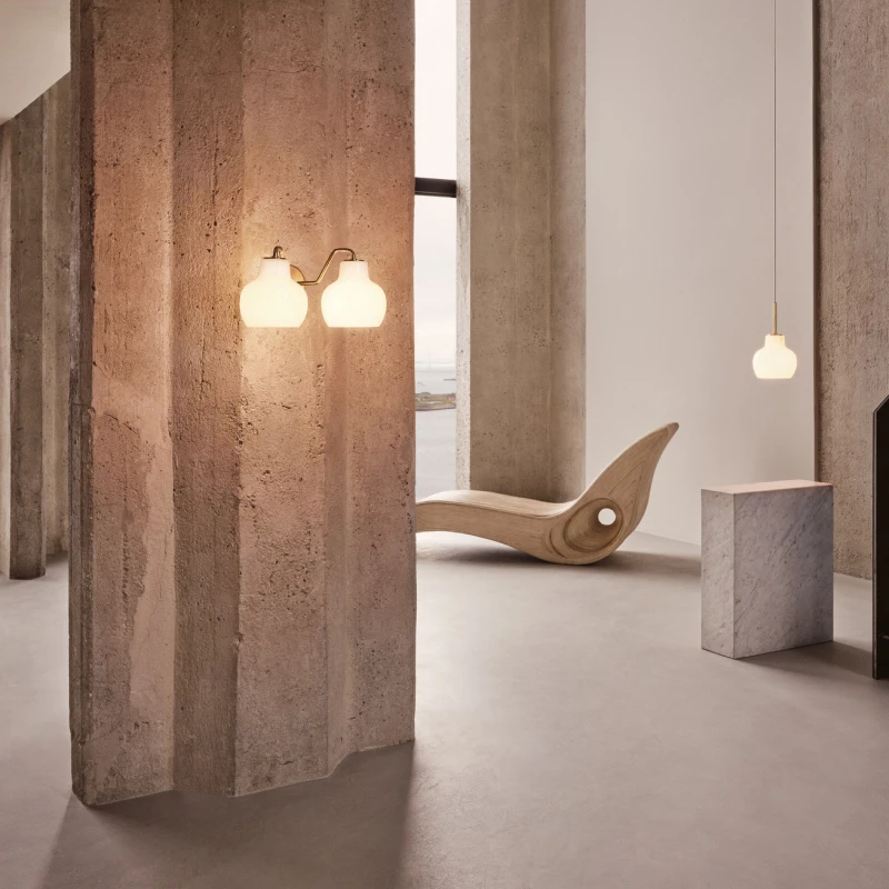 A VL Ring Crown 2 Wall Lamp and VL Ring Crown 1 Pendant glow in a concrete room beside a sculptural lounge chair. © 2026 Louis Poulsen.
