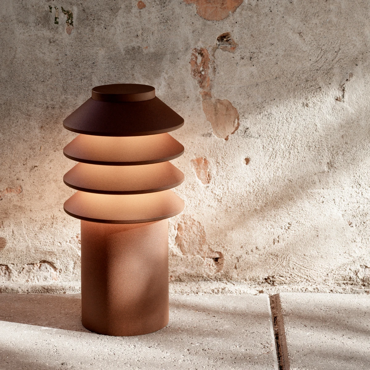 A rust-brown Bysted Garden Bollard glows beside a weathered stone wall and pale paving. © 2026 Louis Poulsen.