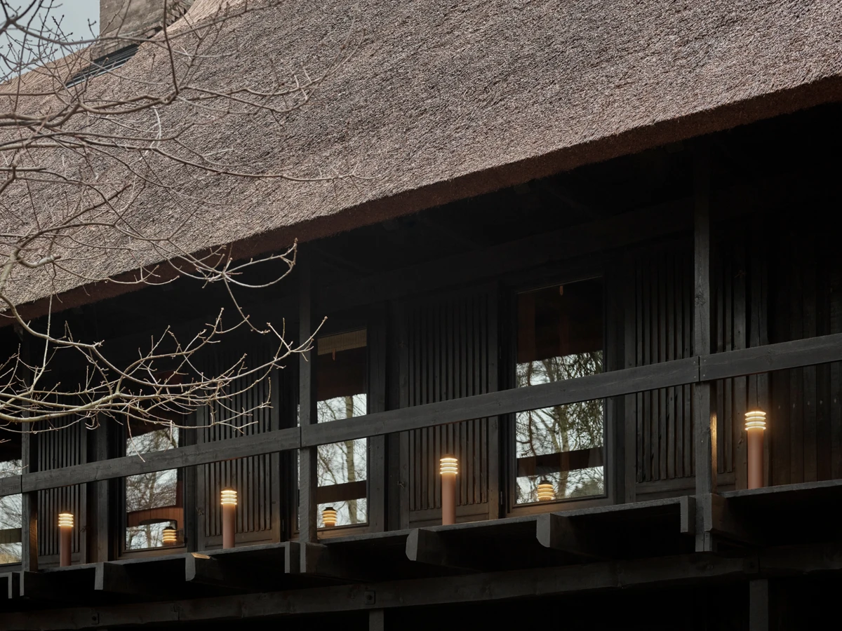 Bysted Garden Bollards illuminate the veranda of a dark timber house with a thatched roof. © 2026 Louis Poulsen.