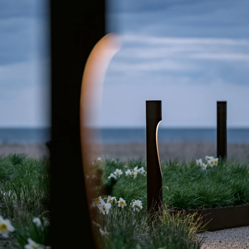 Flindt-bolderarmaturen staan langs een bloembed met siergrassen aan zee in de schemering. © 2026 Louis Poulsen.
