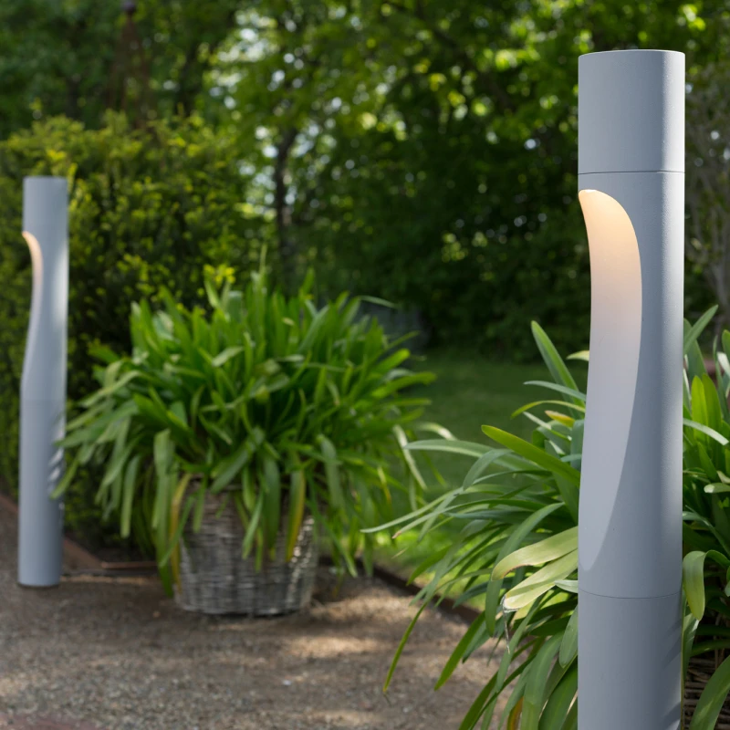 Paletti Flindt grigi diffondono una luce calda lungo un sentiero in giardino, tra una folta vegetazione. © 2026 Louis Poulsen.