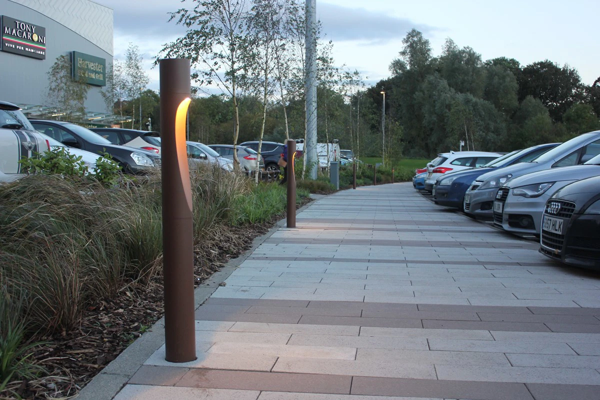 Dark bronze Flindt Bollard lights line a paved walkway beside a car park. © 2026 Louis Poulsen.