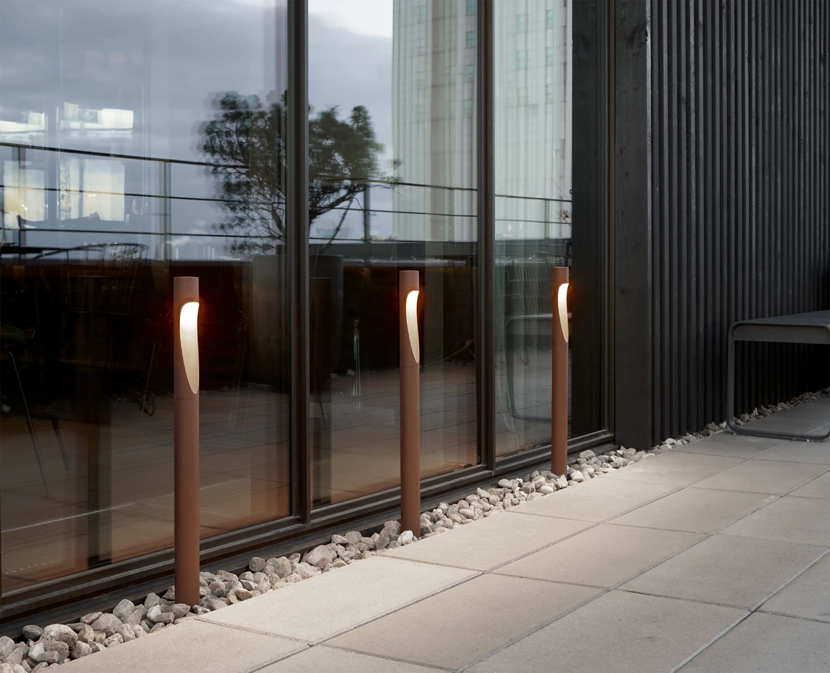 Three brown Flindt Garden Bollards glow beside a paved walkway along a glass facade. © 2026 Louis Poulsen.