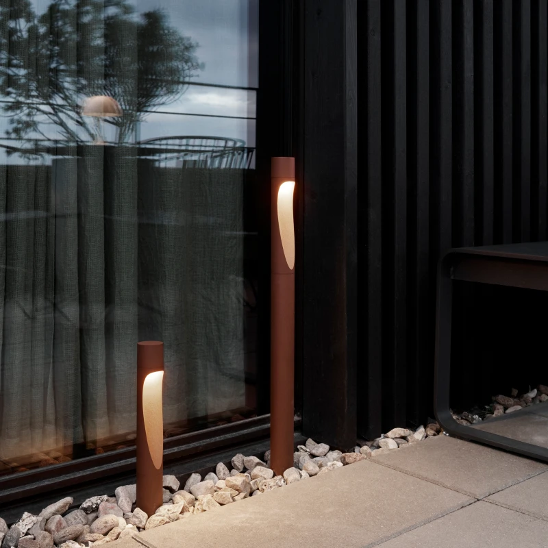 Two brown Flindt Garden Bollards glow beside the glass facade and stone-paved terrace of a modern building. © 2026 Louis Poulsen.