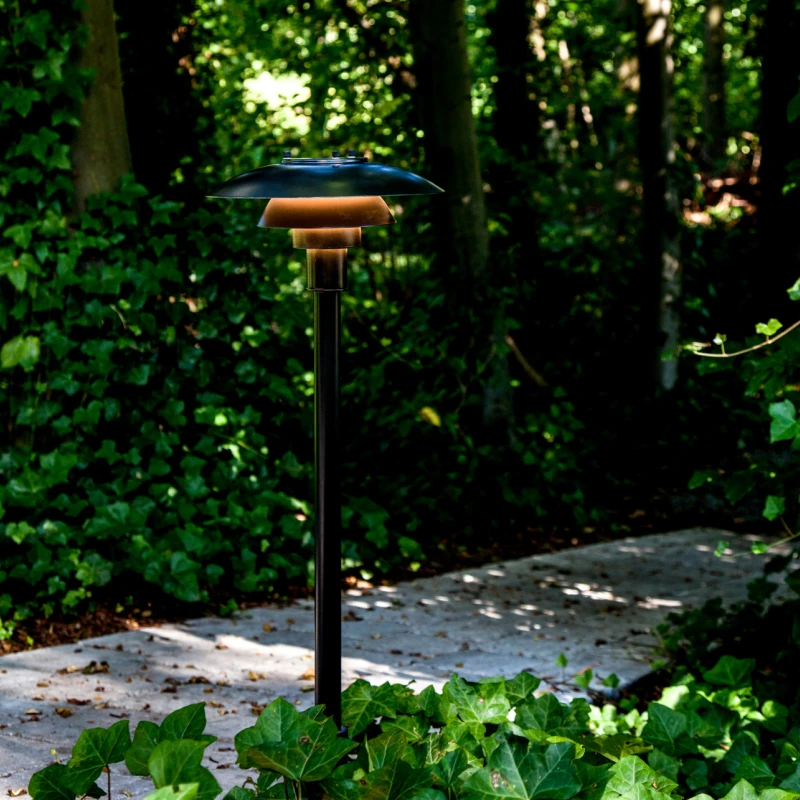 A PH 3-2½ Bollard with a dark shade lights a garden path bordered by dense greenery. © 2026 Louis Poulsen.