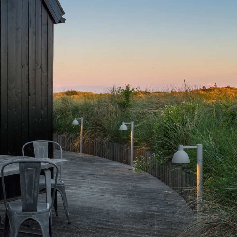 Grey Toldbod 155 Bollards line a wooden deck beside a cabin among the dunes at dusk. © 2026 Louis Poulsen.