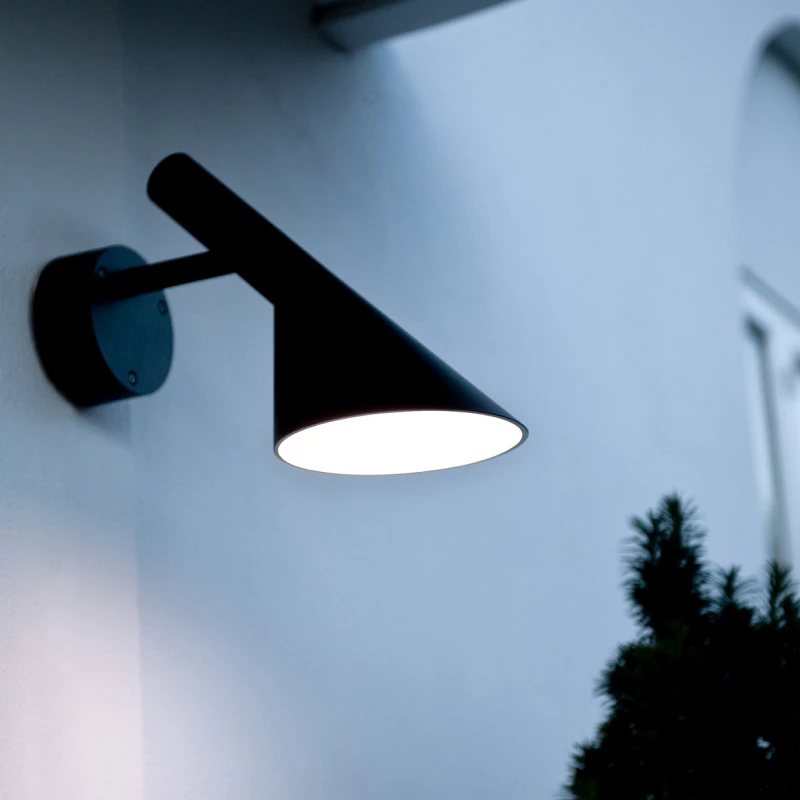 A black AJ 50 Outdoor Wall Lamp illuminates a pale exterior wall at dusk. © 2026 Louis Poulsen.