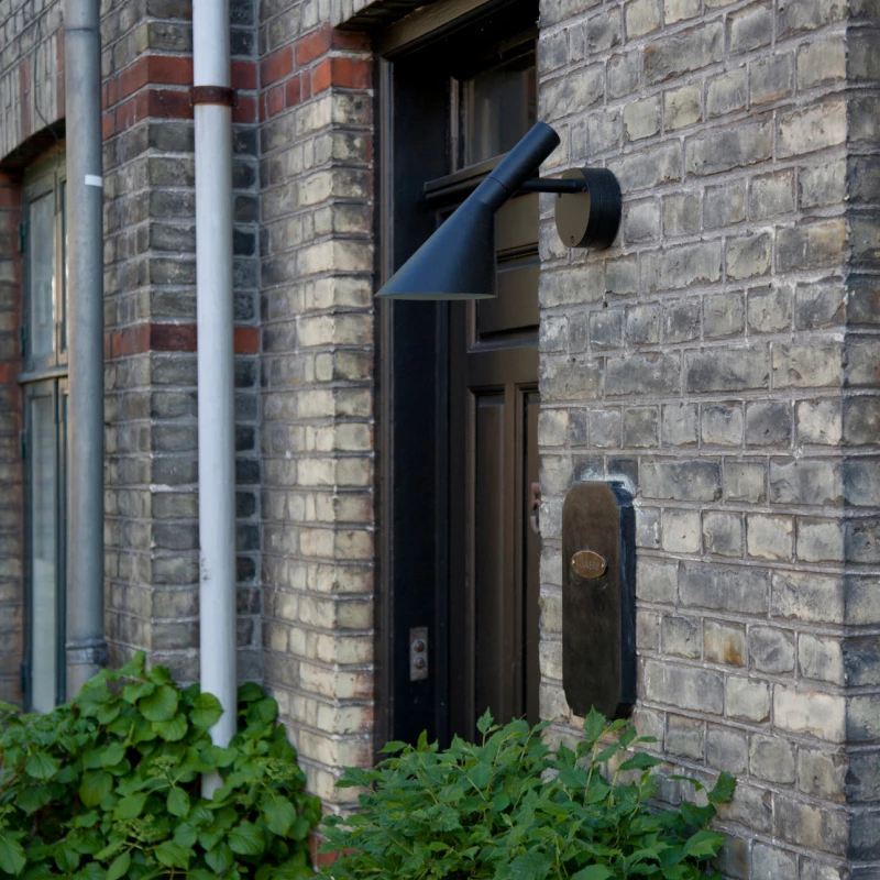 A black AJ 50 Outdoor Wall Lamp on a brick facade beside an entrance framed by greenery. © 2026 Louis Poulsen.