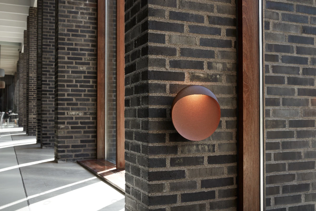 A corten Flindt Wall Lamp casts light on a dark brick wall beside a modern building. © 2026 Louis Poulsen.
