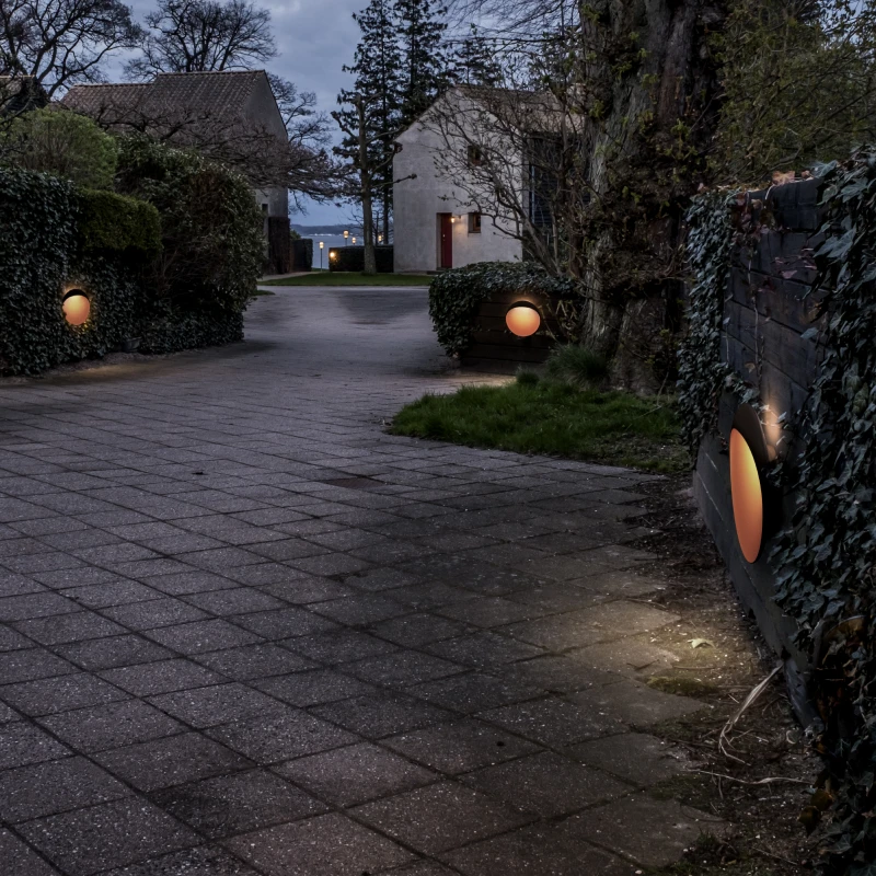 Flindt Wall Lamp wall lights illuminate a paved driveway bordered by hedges and stone walls at dusk. © 2026 Louis Poulsen.