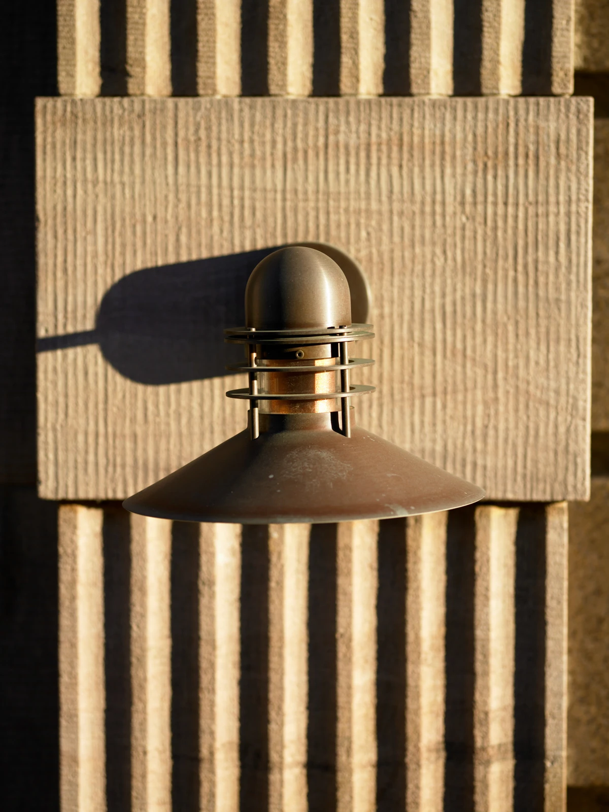 A bronze Nyhavn Outdoor Wall Lamp on a ribbed facade in warm sunlight. © 2026 Louis Poulsen.