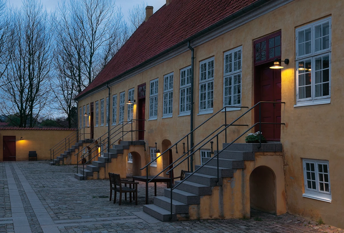 Nyhavn Outdoor Wall Lamps cast a warm glow on a yellow barracks building and its paved courtyard at dusk. © 2026 Louis Poulsen.