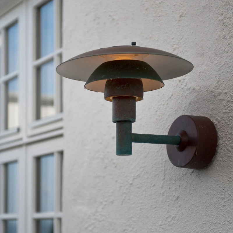 A PH 3-2½ Outdoor Wall Lamp with a dark, patinated finish mounted on a white exterior wall beside a window. © 2026 Louis Poulsen.