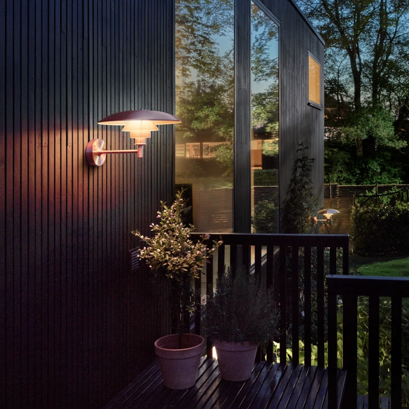 A PH 3-2½ Outdoor Wall Lamp casts a warm glow on a dark timber facade beside a planted deck. © 2026 Louis Poulsen.