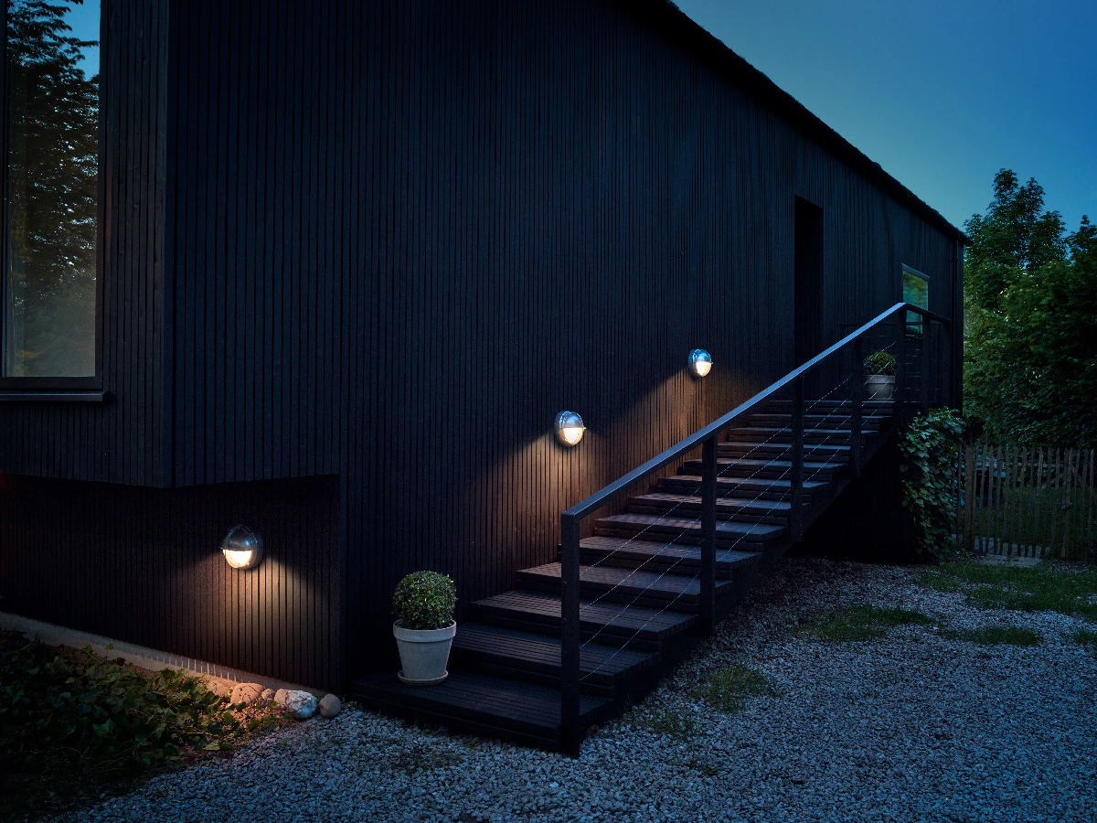 Three Skot Wall Lamp wall lights illuminate a dark timber facade and outdoor stairs in a Birkerød garden. © 2026 Louis Poulsen.