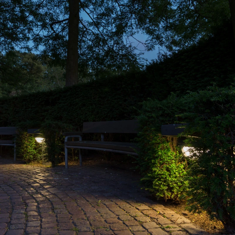 Skot Wall Lamps illuminate a cobbled path between benches and dense greenery at dusk. © 2026 Louis Poulsen.