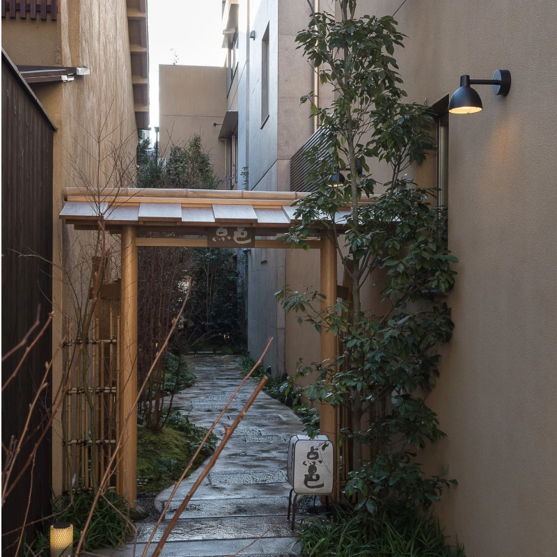 A black Toldbod 155 Outdoor Wall Lamp illuminates a narrow garden path beside a wooden entrance. © 2026 Louis Poulsen.