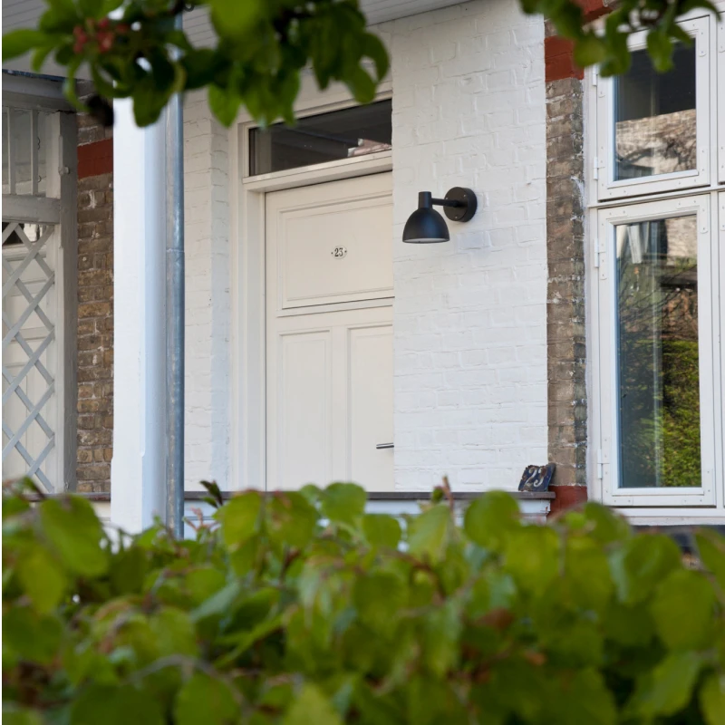 A black Toldbod 155 Outdoor Wall Lamp beside a white front door on a brick facade, framed by green foliage. © 2026 Louis Poulsen.
