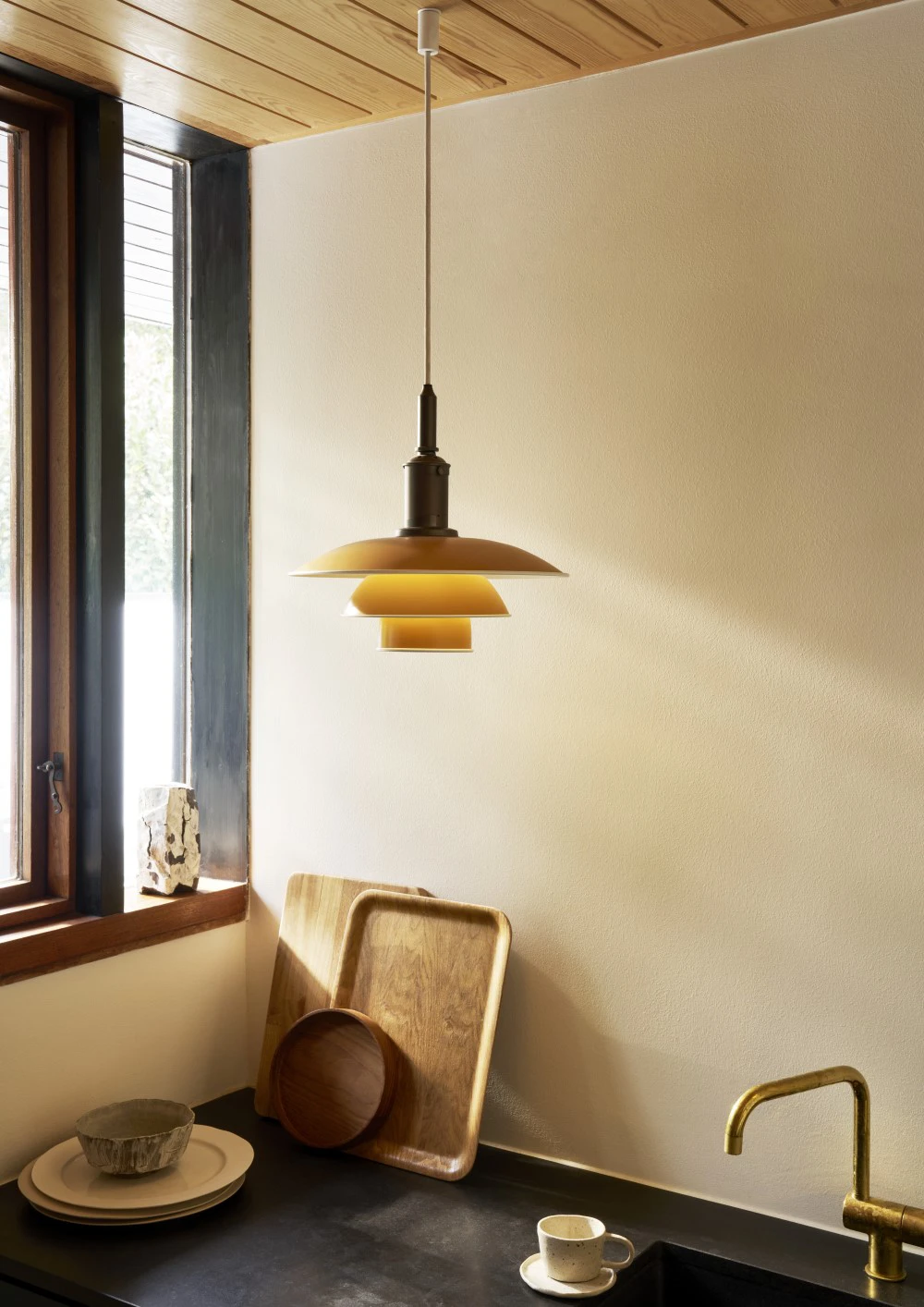 A PH 3½-3 Amber Coloured Pendant casts a warm glow over a kitchen counter beside a window. © 2026 Louis Poulsen.