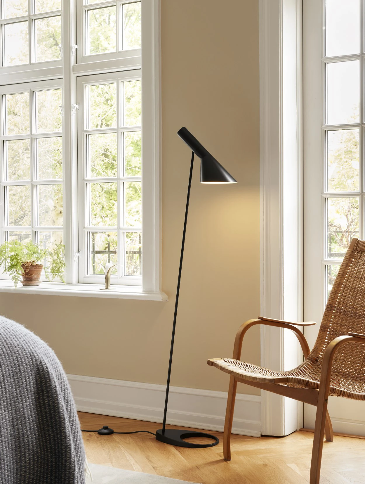 A black AJ Floor Lamp illuminates a wicker chair in a bright living room with tall windows and wooden floors. © 2026 Louis Poulsen.