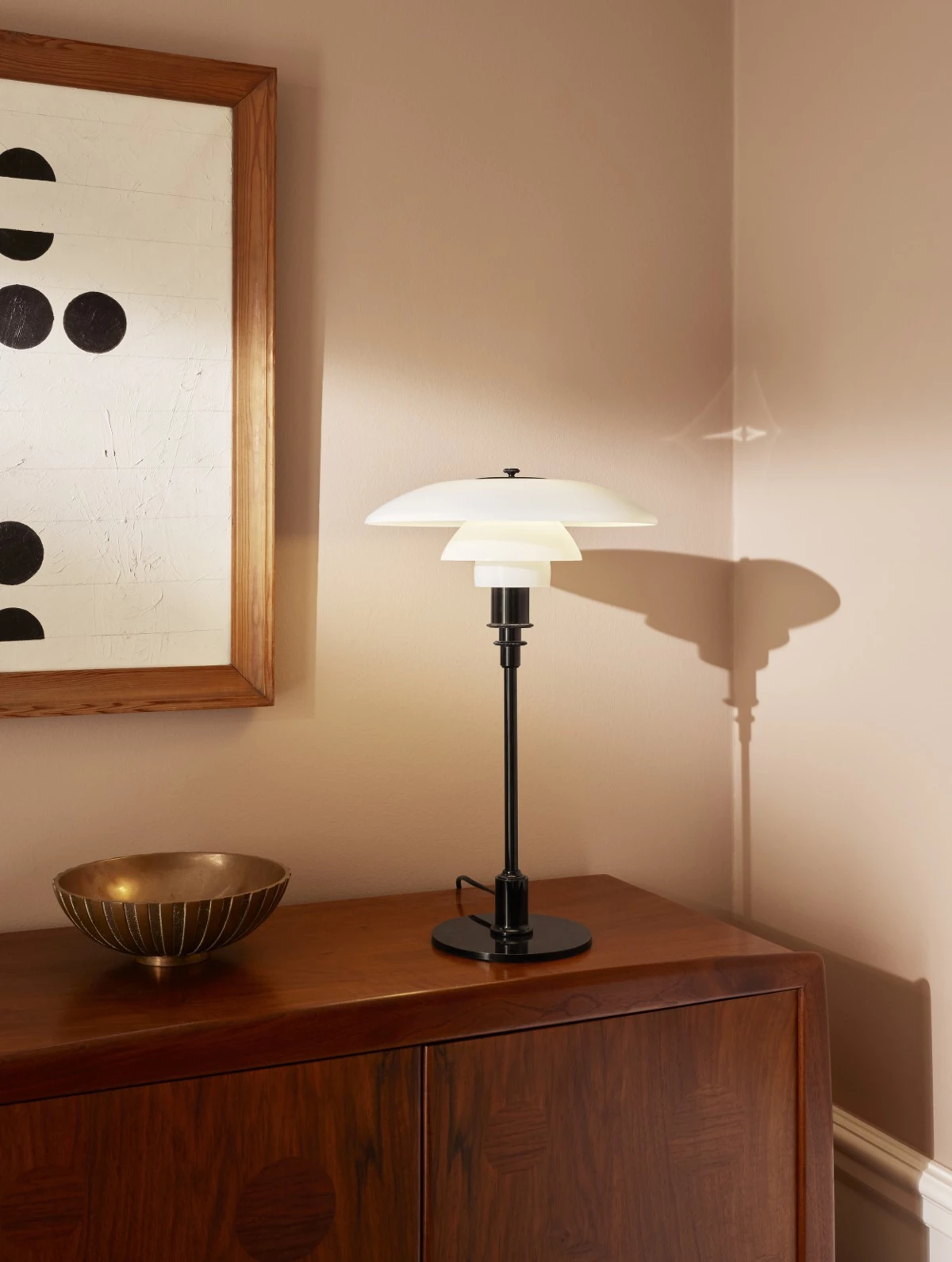 A PH 3½-2½ Table Lamp with white shades and a black stem sits on a wooden cabinet beside framed artwork. © 2026 Louis Poulsen.