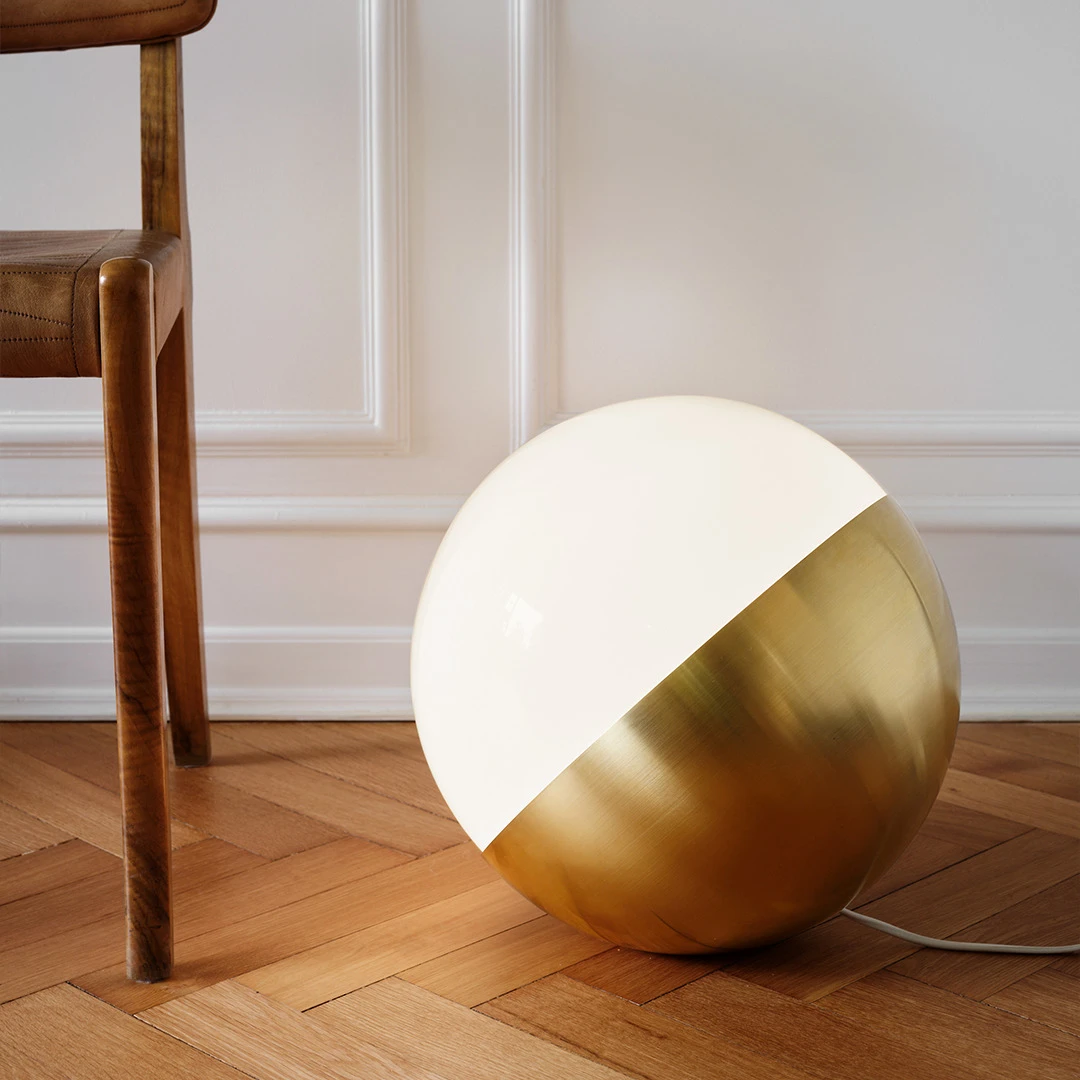 The brass VL Studio Table/Floor Lamp with an opal-white shade sits beside a leather chair on a herringbone floor. © 2026 Louis Poulsen.
