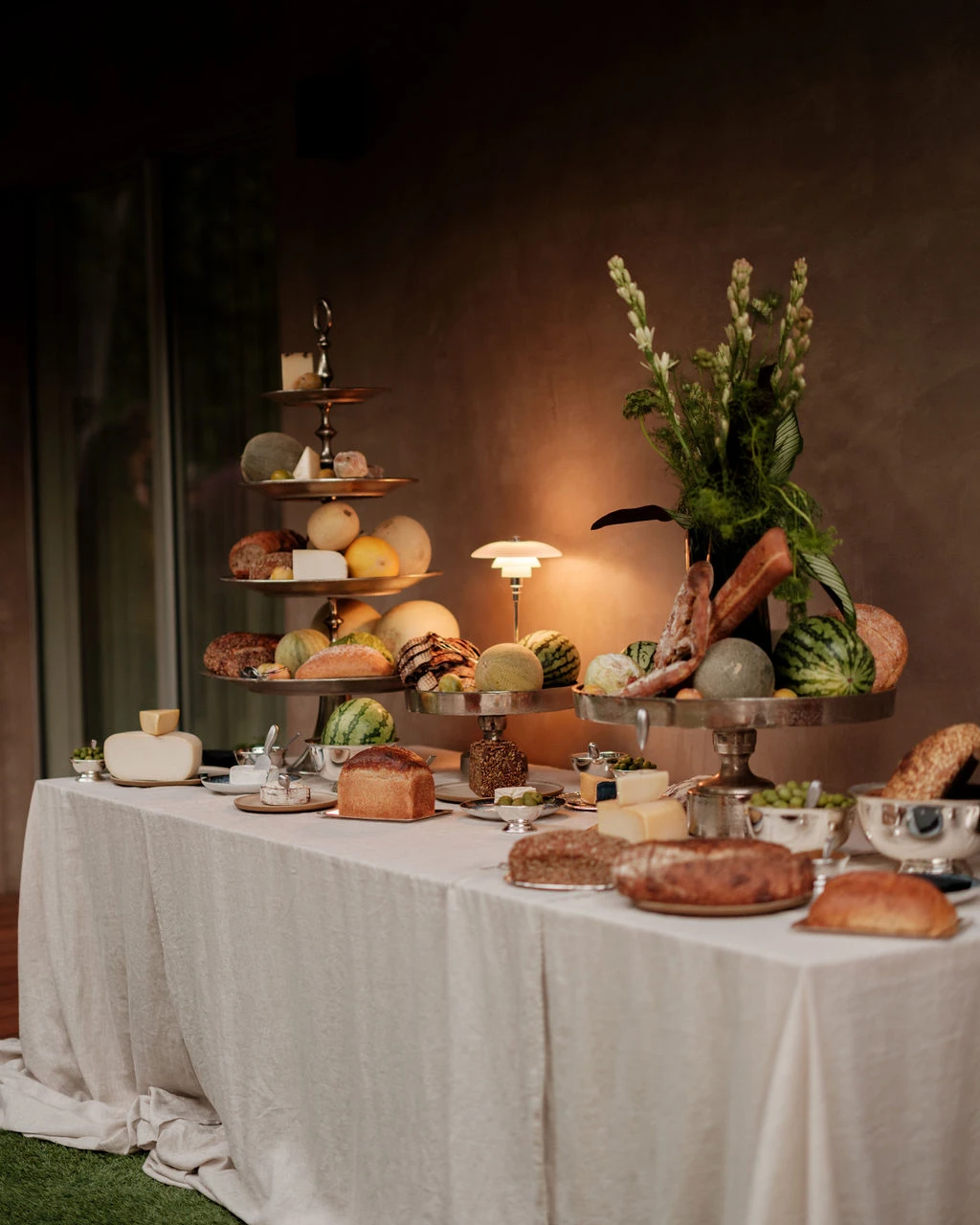 A PH 2/1 Table Lamp casts a warm glow over a buffet table laden with bread, fruit and flowers. © 2026 Louis Poulsen.