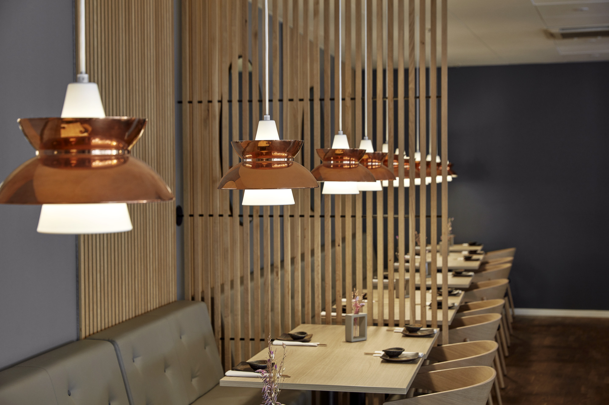 Copper-coloured Doo-Wop pendants hang above dining tables in a sushi restaurant with timber slat partitions. © 2026 Louis Poulsen.