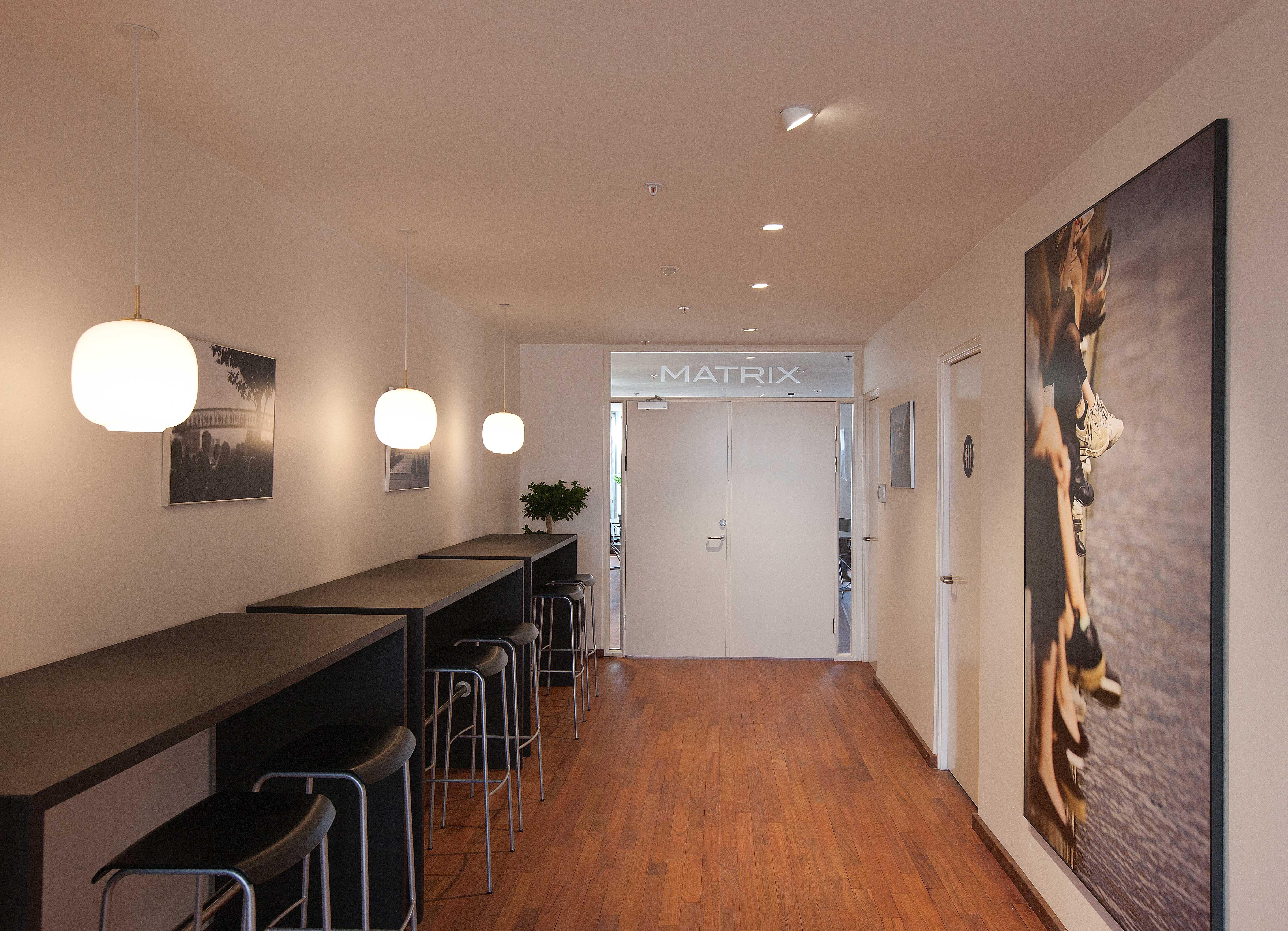 Three white Wohlert Pendants light a hallway with wooden flooring, high counters and bar stools. © 2026 Louis Poulsen.