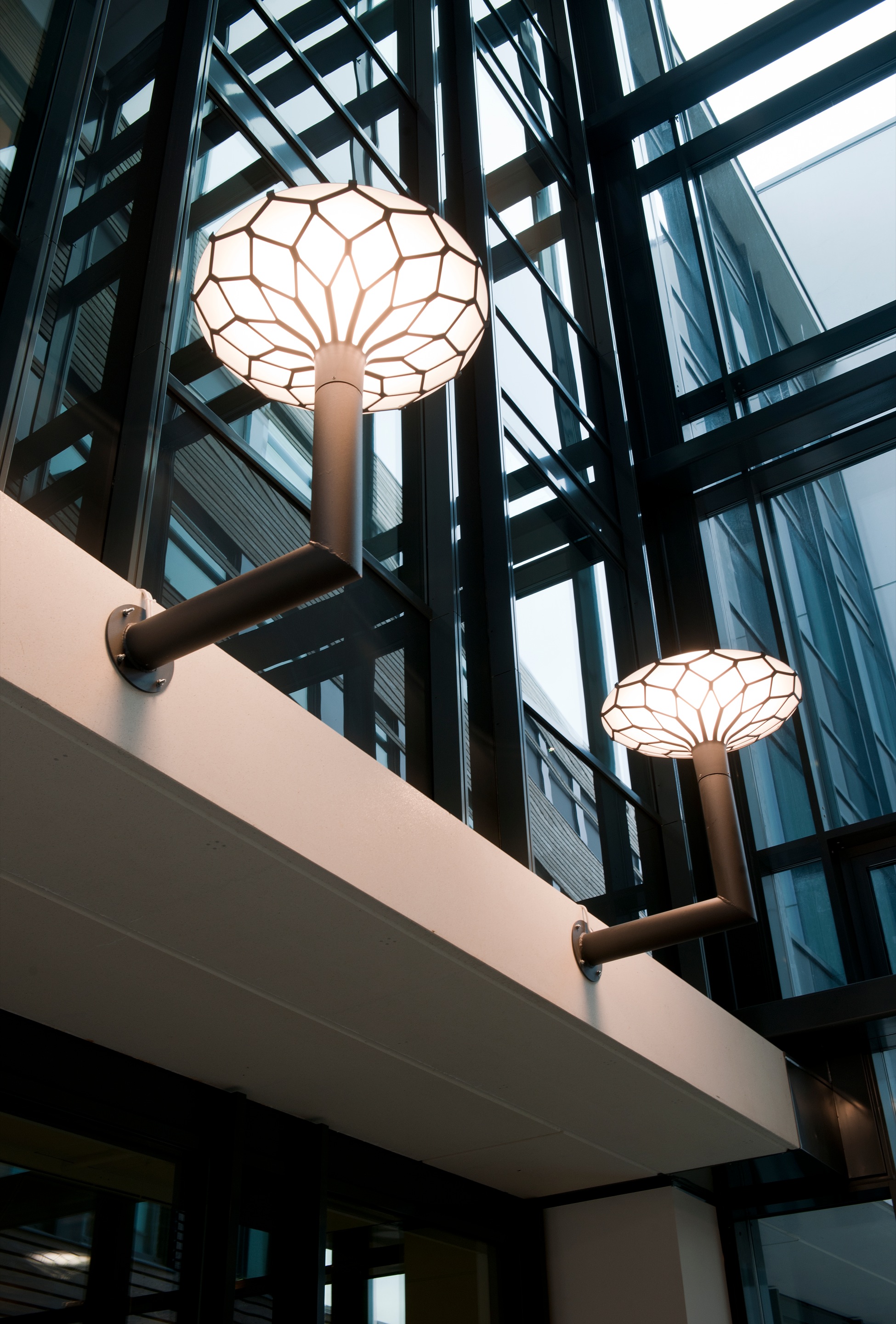 Two opal-white Louis Poulsen wall lamps illuminate a modern atrium with tall glass façades and dark steel beams. © 2026 Louis Poulsen.