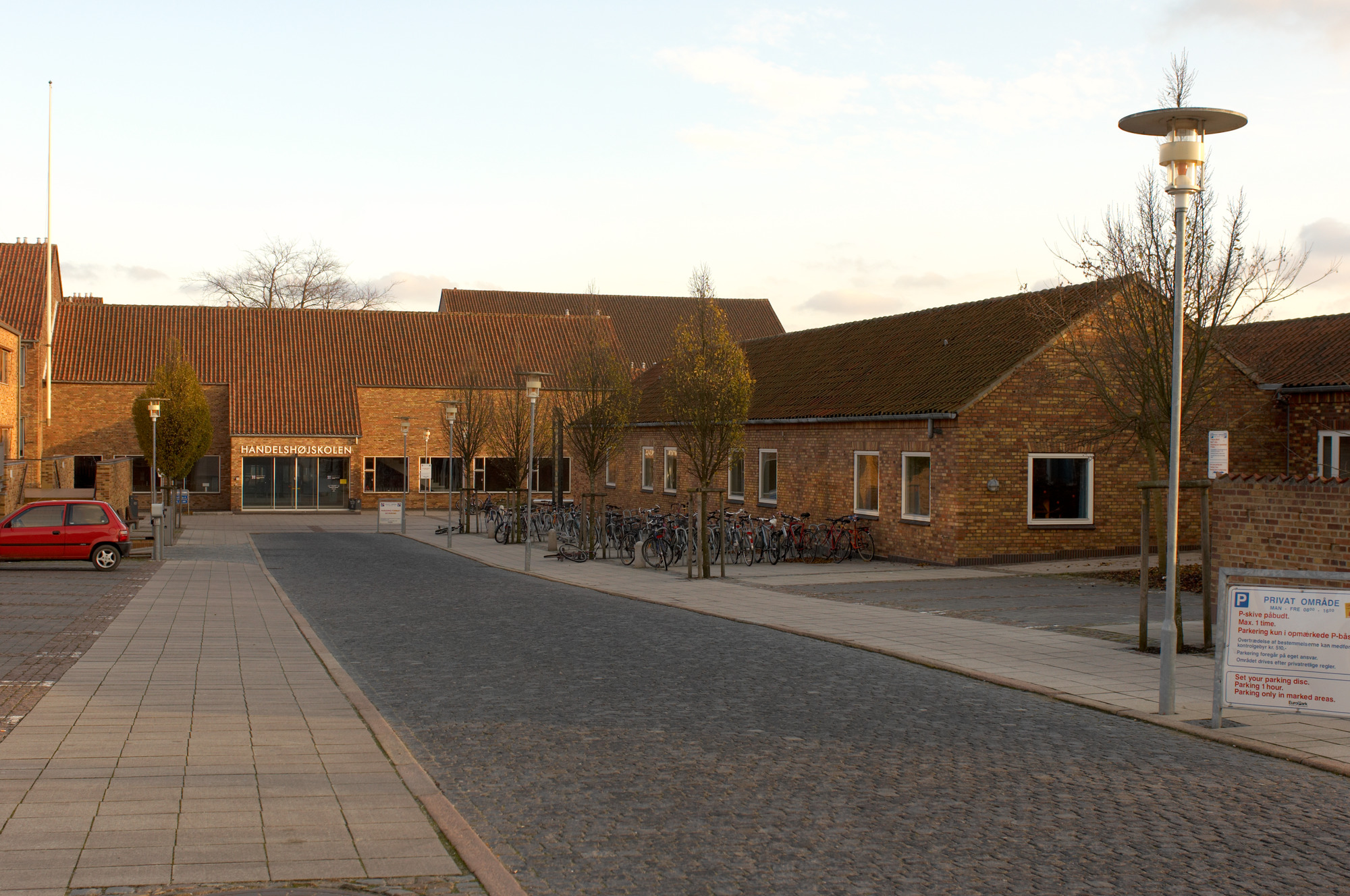 An Albertslund Maxi Post stands beside a cobbled road in front of the low brick buildings of Aarhus Handelshøjskole. © 2026 Louis Poulsen.