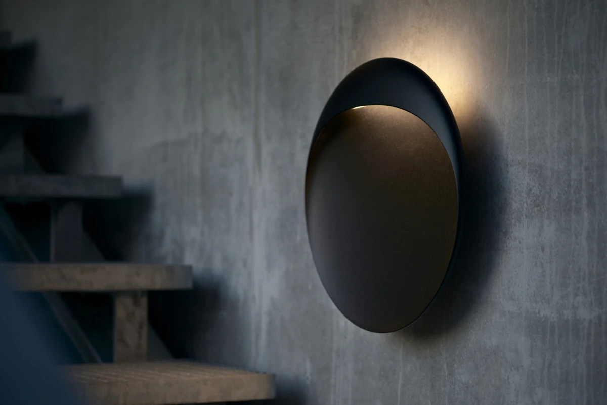 Une applique Flindt noire diffuse une lumière chaude sur un mur en béton gris près d’un escalier. © 2026 Louis Poulsen.