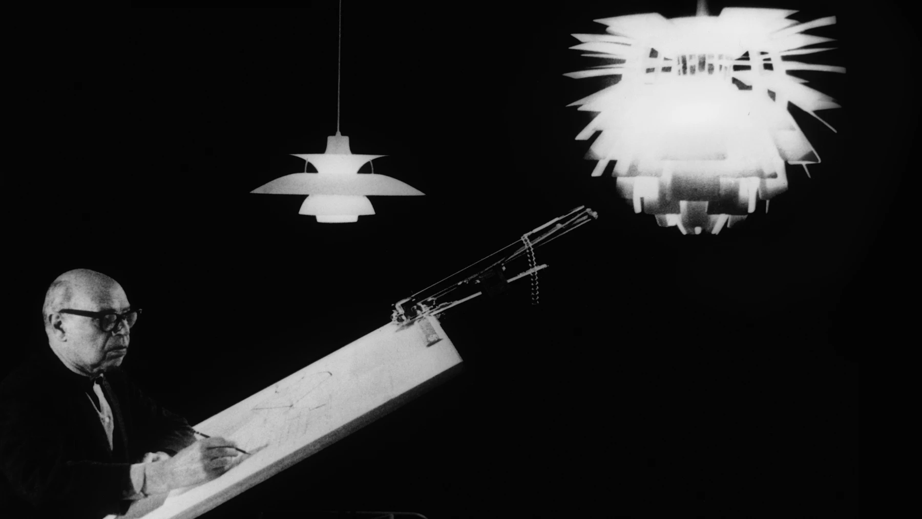 A PH 5 pendant and PH Artichoke pendant hang above a designer at a drafting table in a black-and-white studio photograph. © 2026 Louis Poulsen.