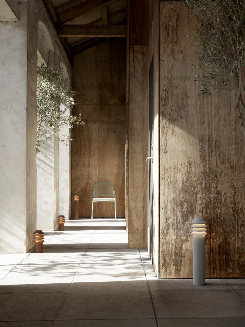 Copper-coloured and grey Bysted Garden Bollard bollards line a sunlit walkway between rustic courtyard walls. © 2026 Louis Poulsen.