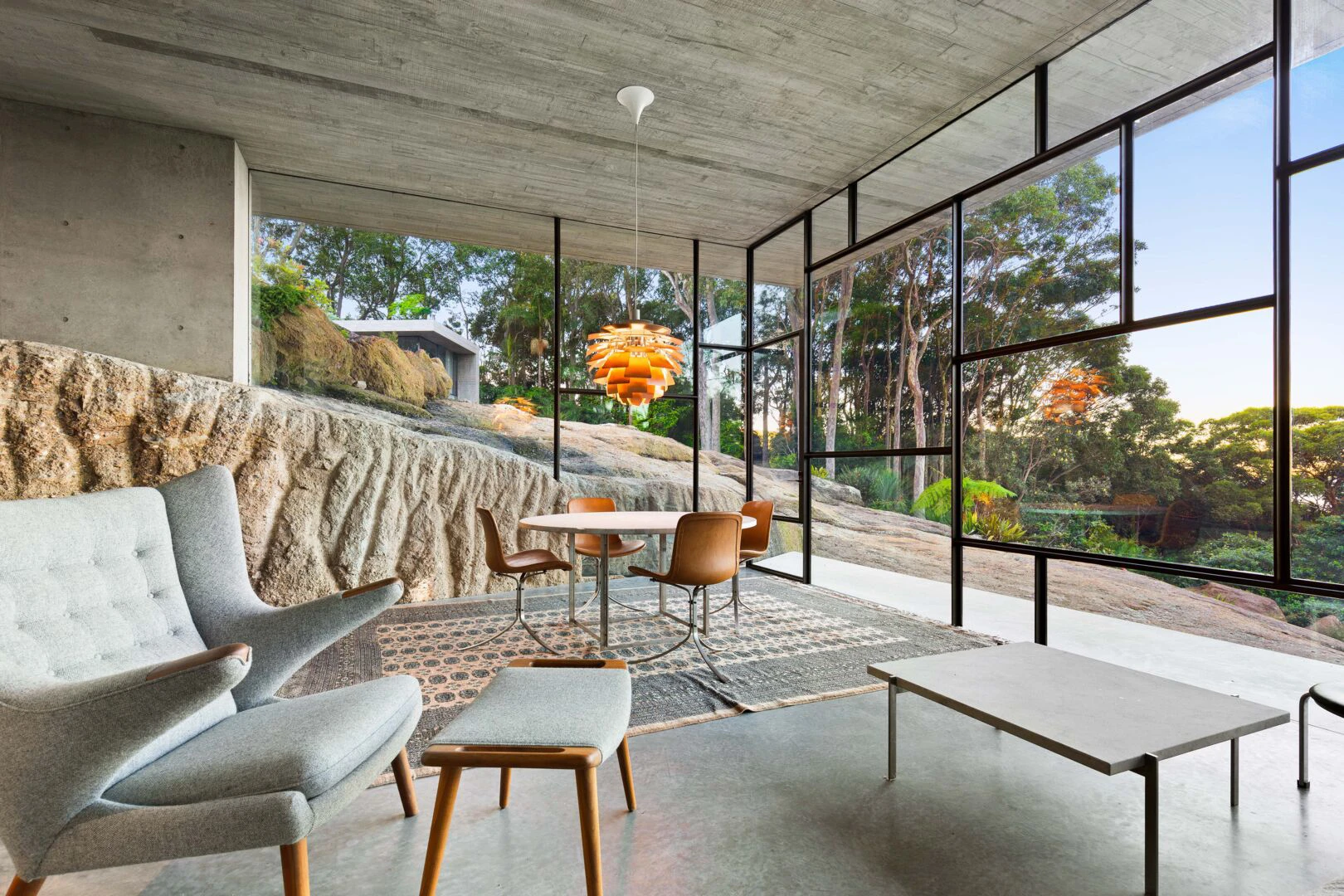 A copper-toned PH Artichoke pendant hangs above the dining table in a glass-walled home overlooking rocks and trees. © 2026 Louis Poulsen.