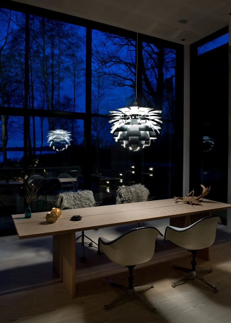 A white PH Artichoke pendant hangs above a long wooden table in a glass-walled room at dusk. © 2026 Louis Poulsen.