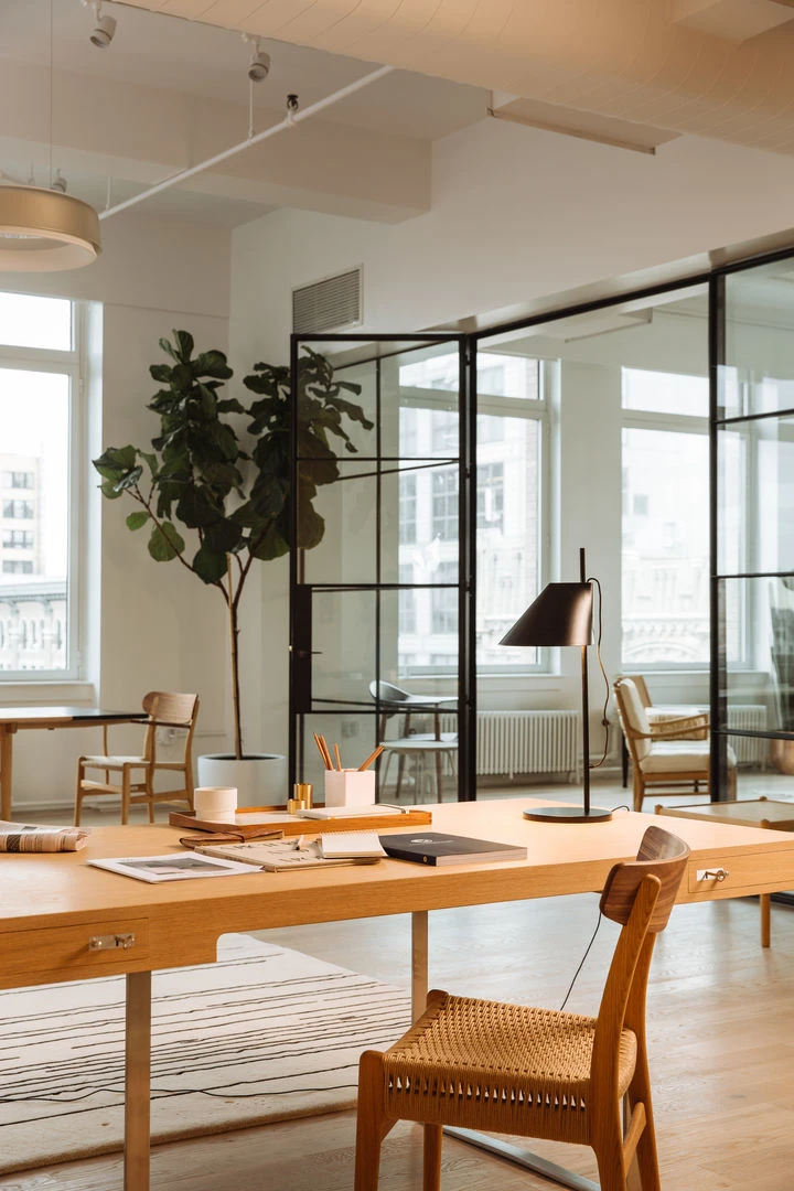 A dark AJ Table Lamp on a light wooden desk in an open office with glass partitions and large windows. © 2026 Louis Poulsen.