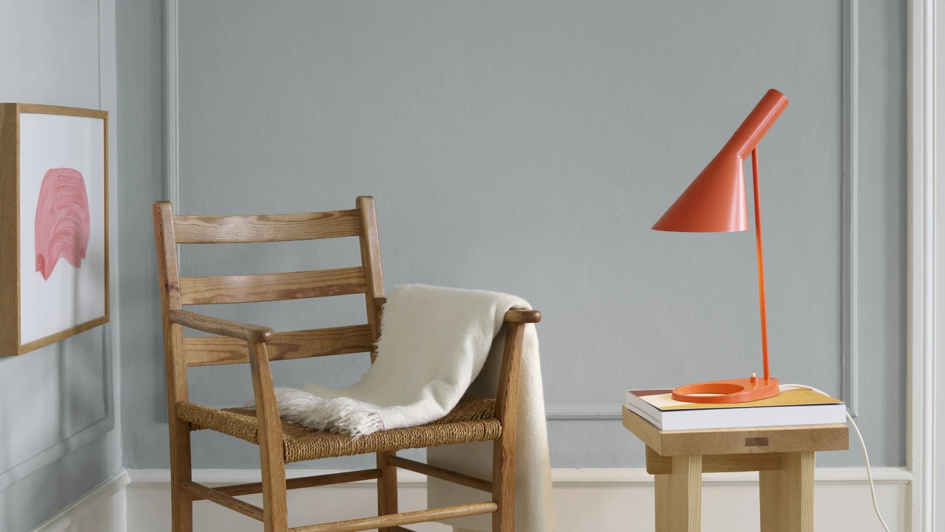 An orange AJ Table Lamp sits on a small wooden table beside an armchair in a softly lit room. © 2026 Louis Poulsen.