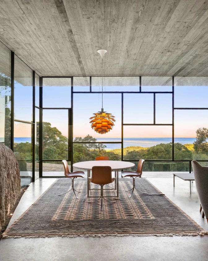 Een oranje PH Artichoke hanglamp hangt boven de eettafel in een glazen ruimte met uitzicht op zee. © 2026 Louis Poulsen.