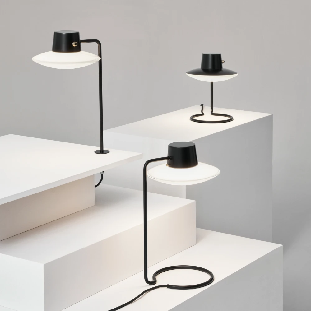 Three black Ballerup table lamps with white shades stand on white plinths against a light grey background. © 2026 Louis Poulsen.